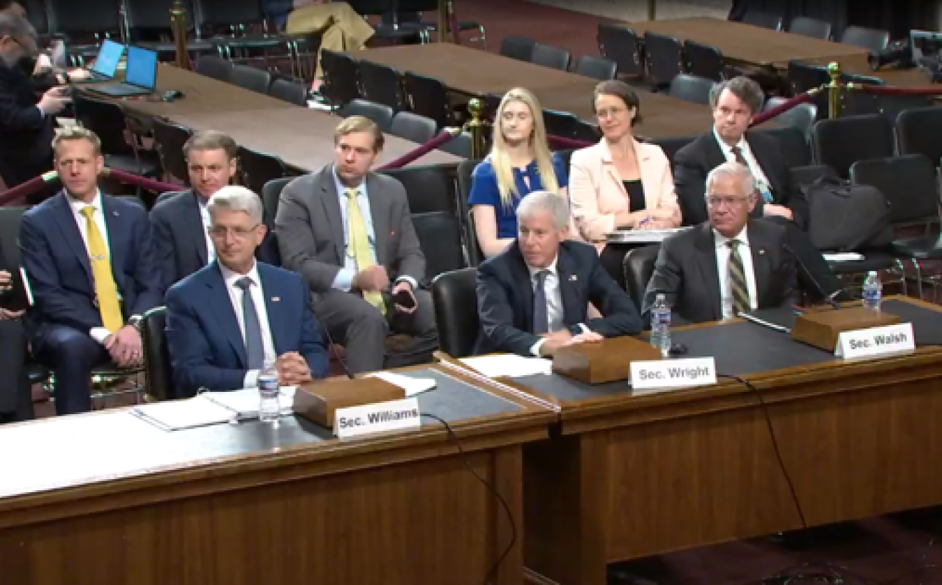 Three men in business suits look to the left of the photo. Behind them are a row of six people also looking to the left. From left, name tags say Sec. Williams., Sec. Wright, and Sec. Walsh.