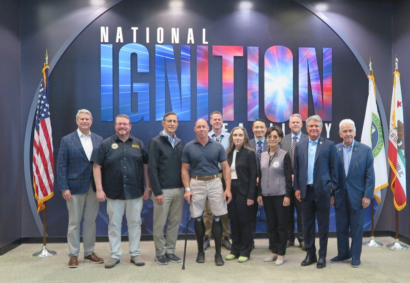 A group of men and womenn stand in front of flags and a large National Ignition Facility logo.