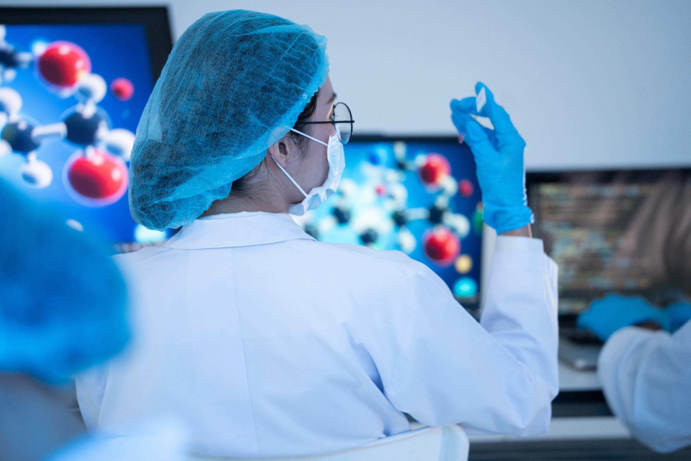 Laboratory researcher wearing protective gear examines a small sample, with molecular models displayed on computer screens.