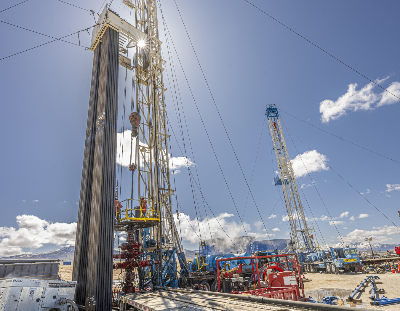 Close-up photo of a drill rig stretching up into a blue sky with the sun shining right behind the derrick (i.e. the tower).