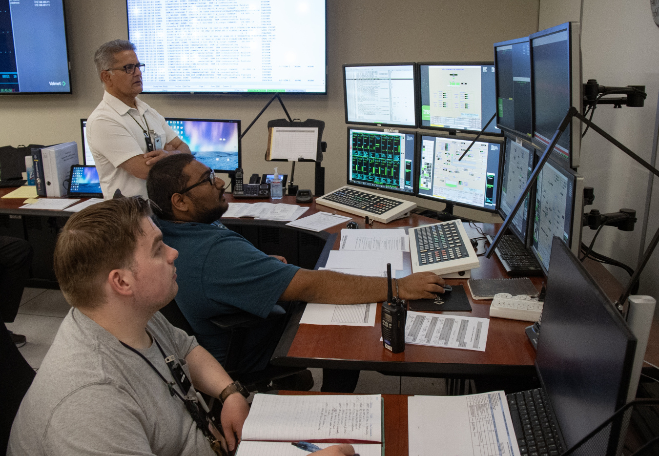 A group of three employees sitting and standing by desks with lots of computer screens, keyboards and papers