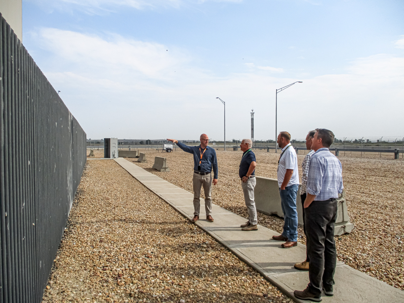 A group of professionals touring Fort St Vrain standing beside a fence on a sidewalk, one man points at the fence