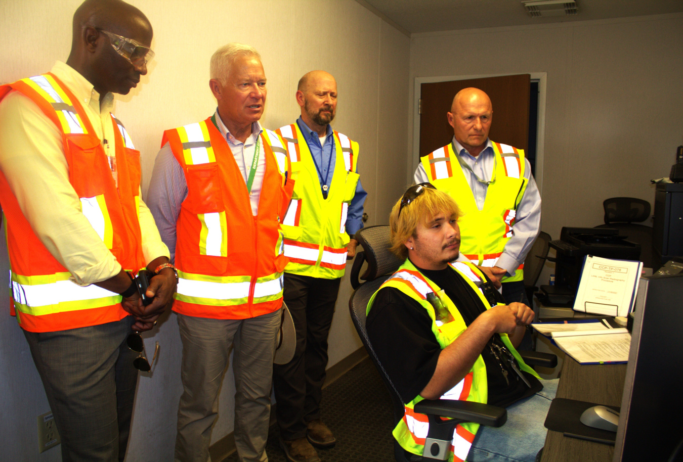One employee sits at a desk in a control room while four other individuals in yellow and orange safety vests stand behind him