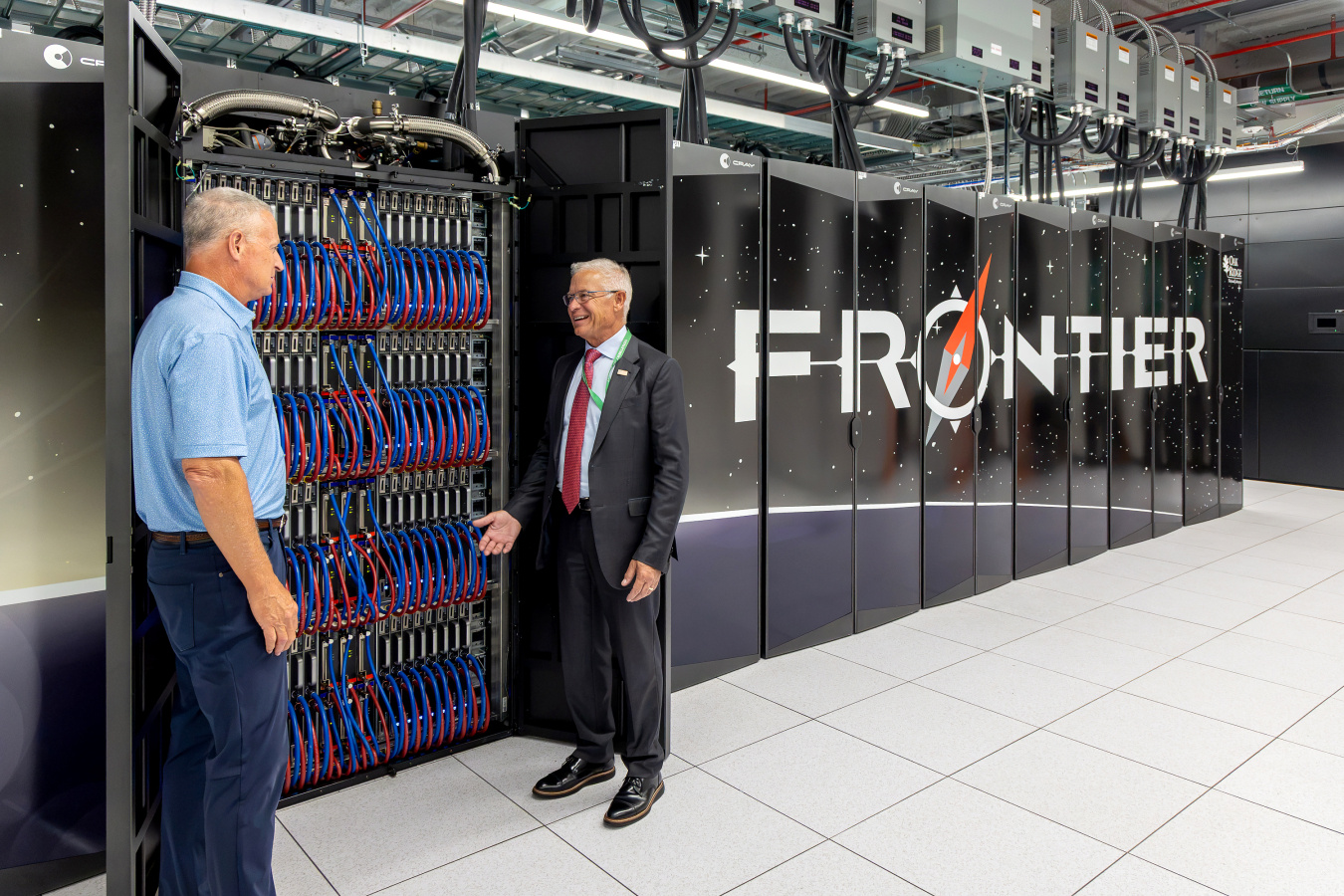 Two professionals speaking with each other in front of a large supercomputer with the words Frontier on it