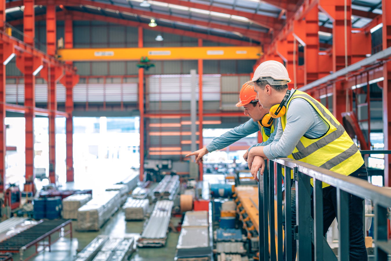 Two workers wearing hard hats and safety vests overlook operations inside a large manufacturing facility. 