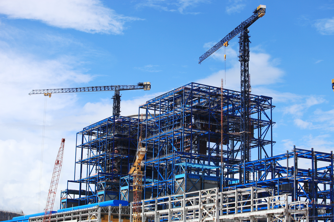 Large steel-frame building under construction with tower cranes against a blue sky. 