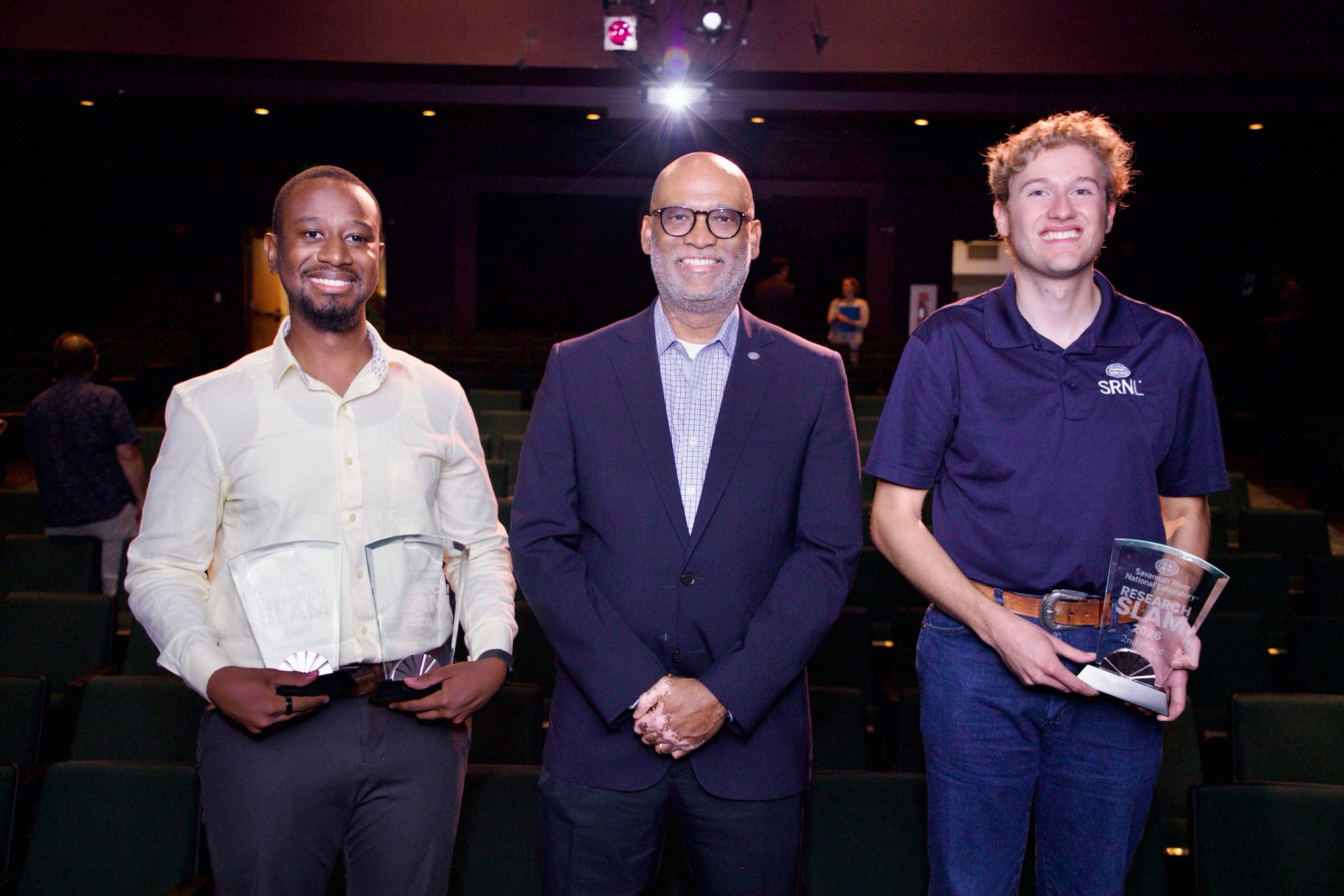 Three professionals pose for a photo and hold clear award plaques