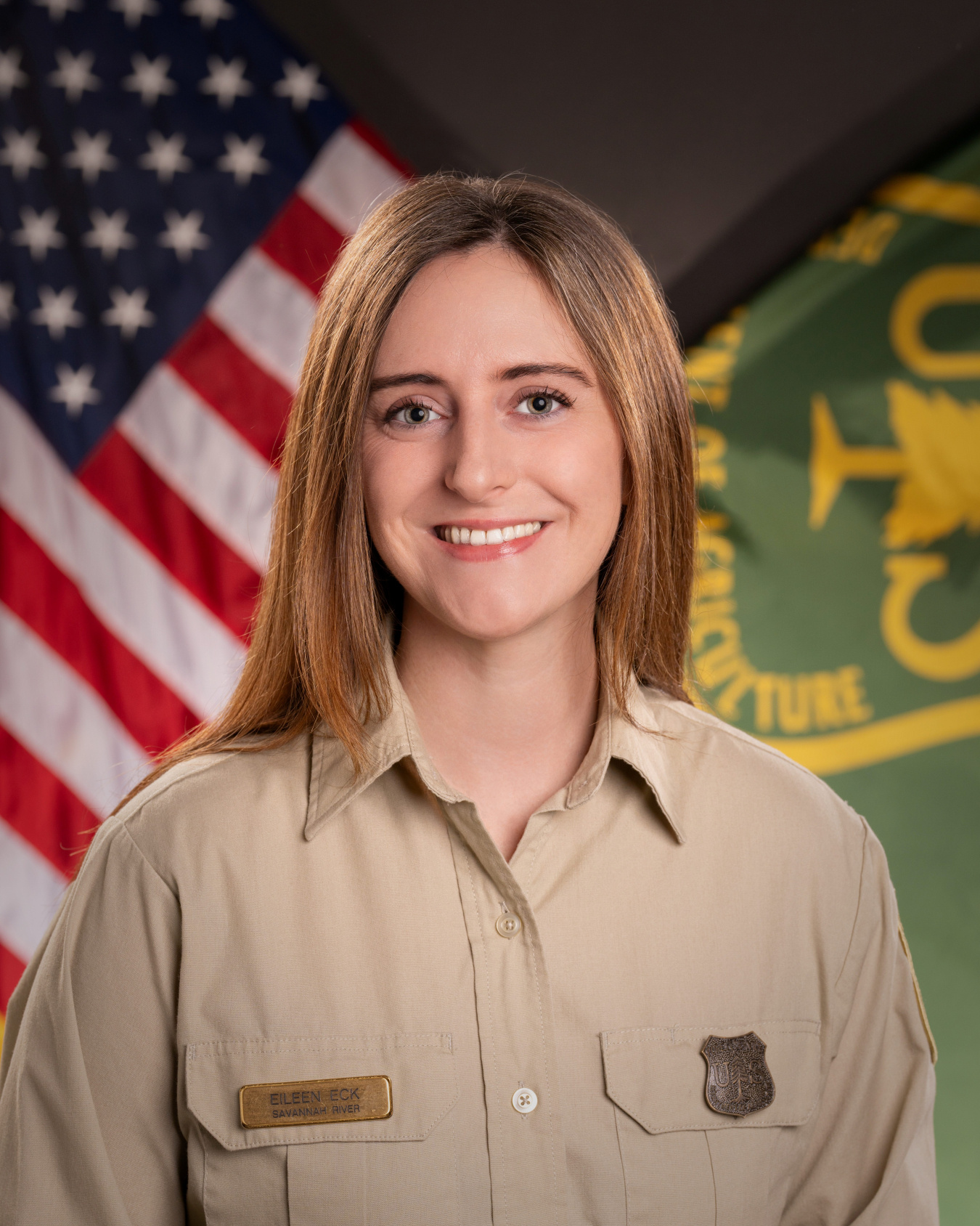 A professional headshot of a Forest Service fellow in a park ranger outfit with an American flag and Forest Service flag in the background