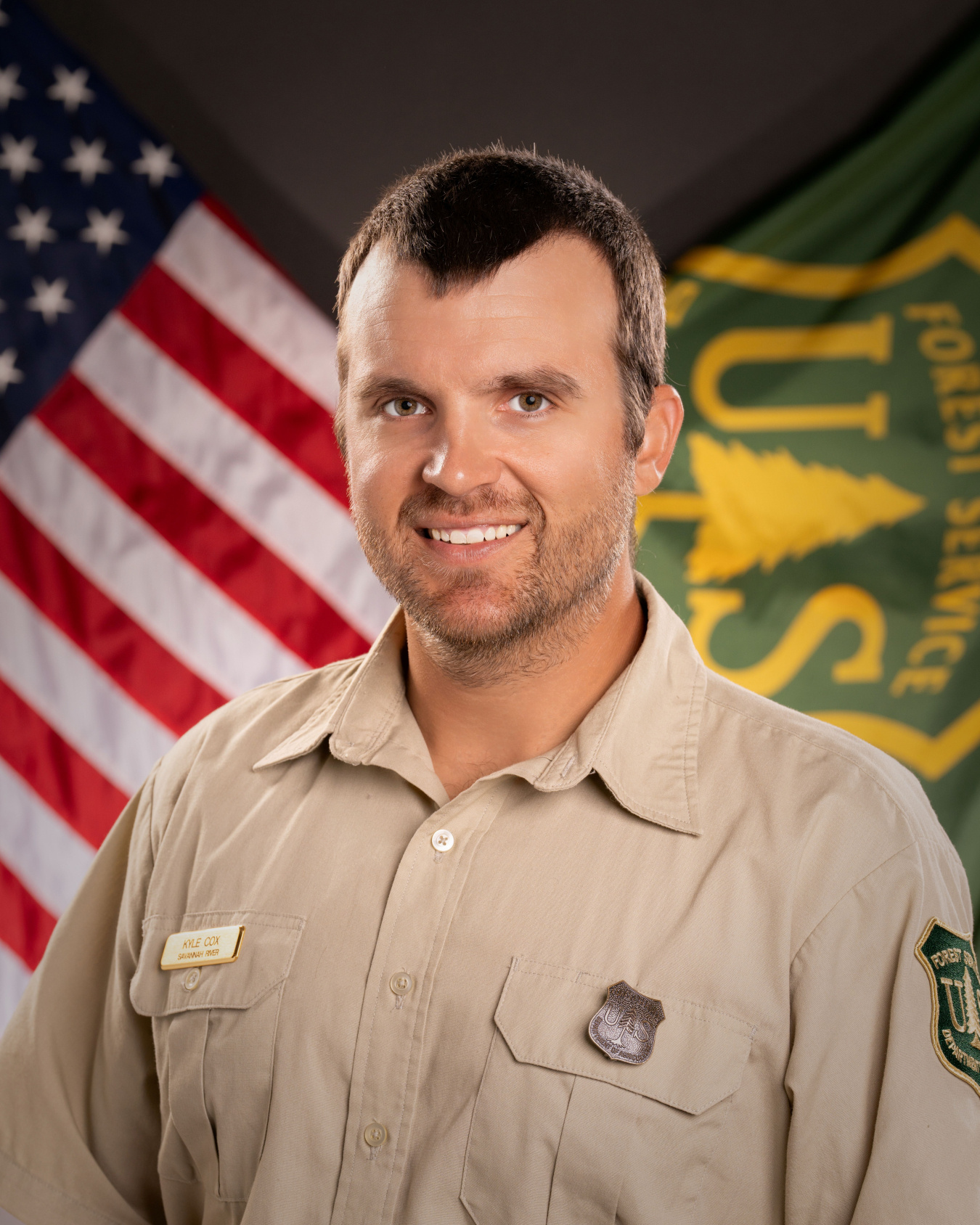 A professional headshot of a Forest Service fellow in a park ranger outfit with an American flag and Forest Service flag in the background