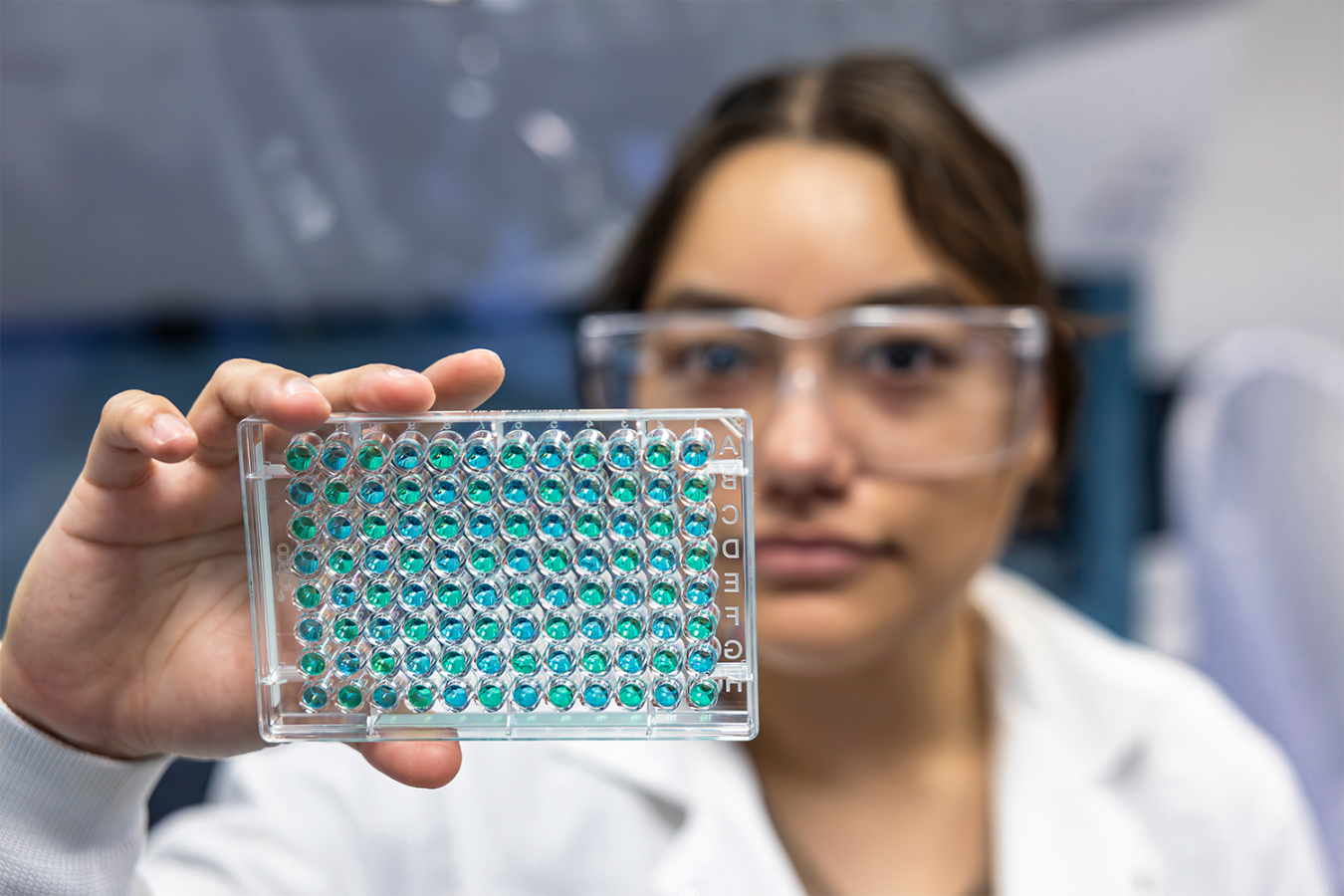 Laboratory researcher wearing safety glasses holds a multiwell plate containing blue and green samples.