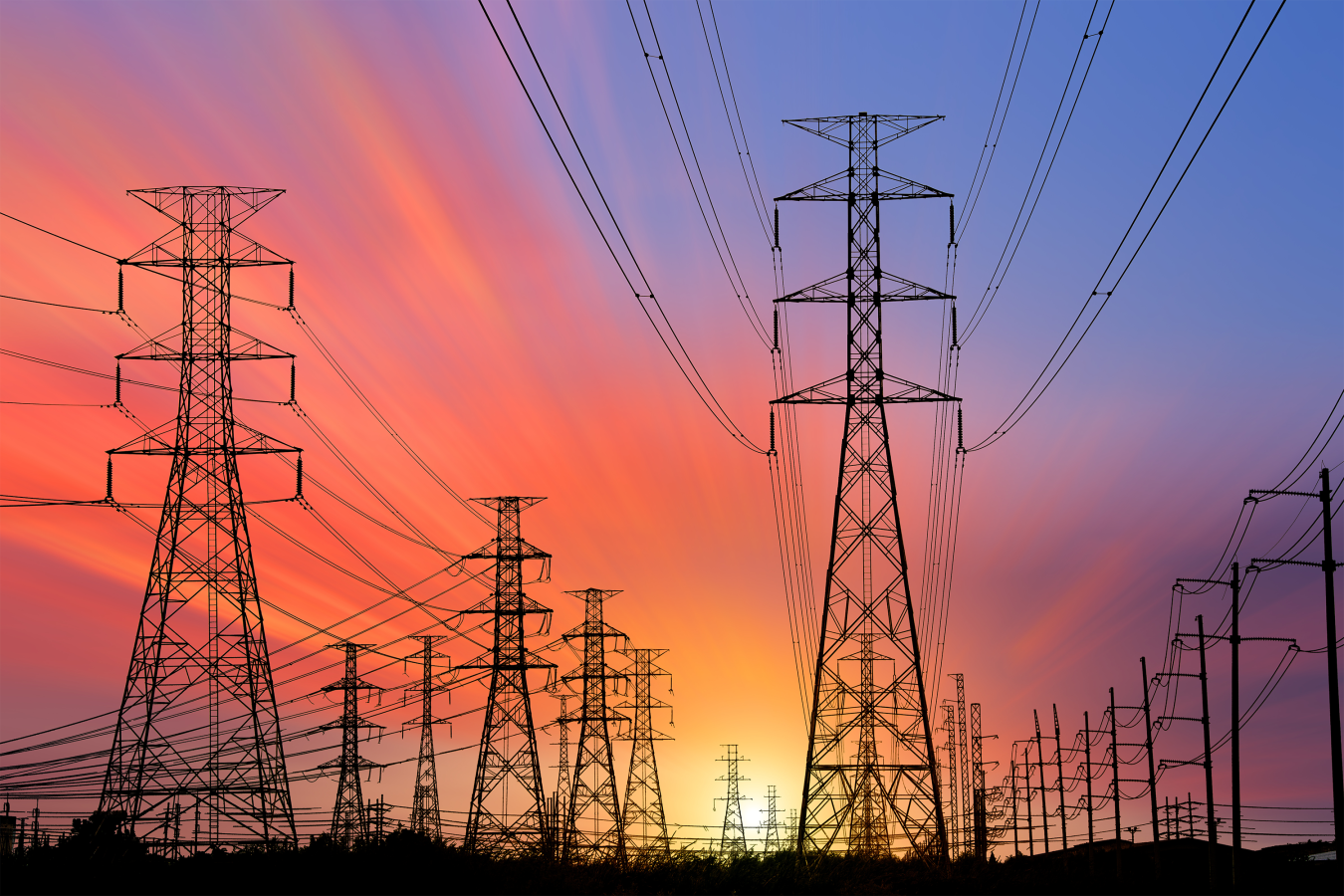 High-voltage electrical transmission towers and power lines silhouetted against a colorful sunset. 