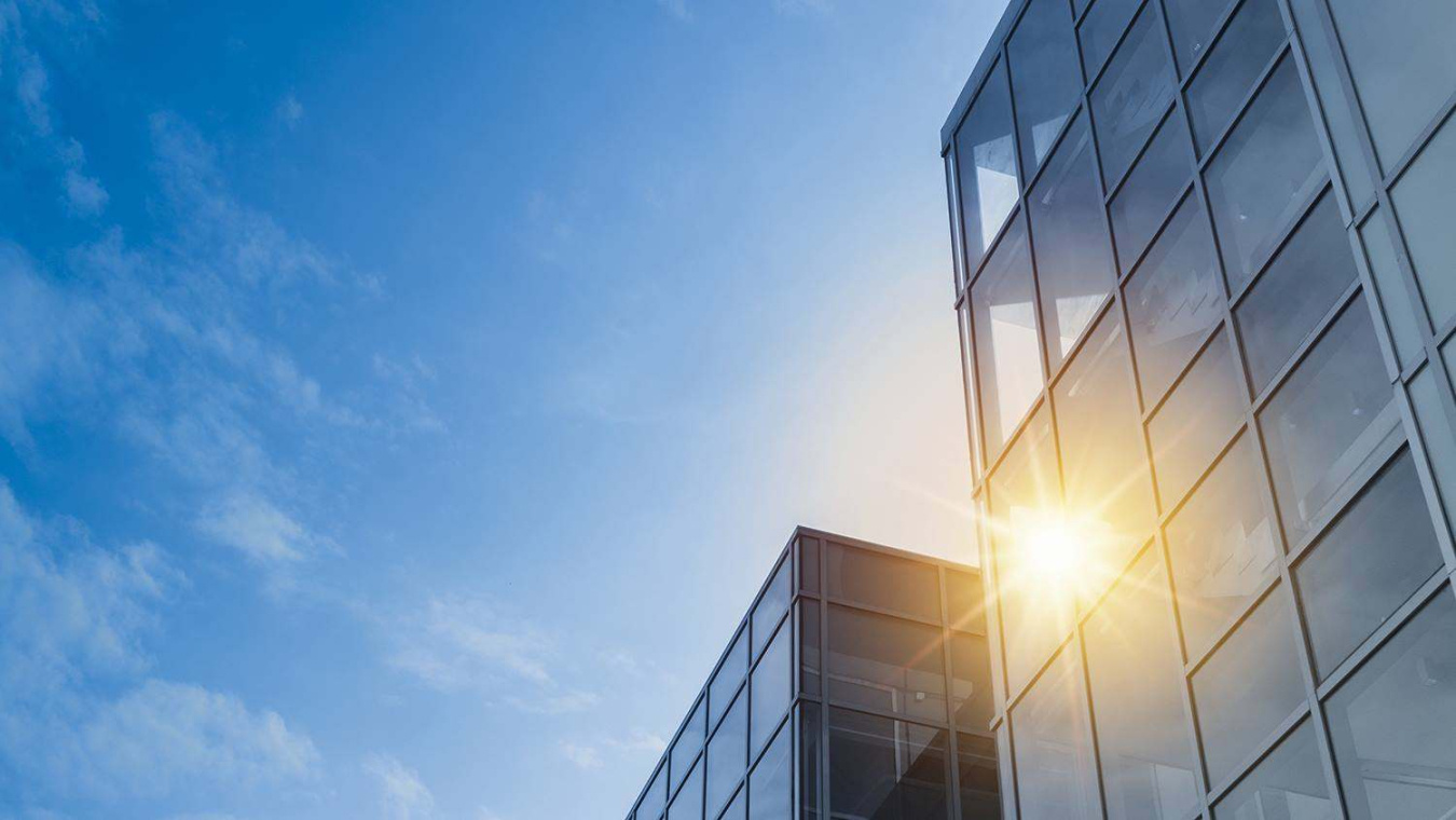 Sunlight shines through the glass facade of a modern building beneath a blue sky.