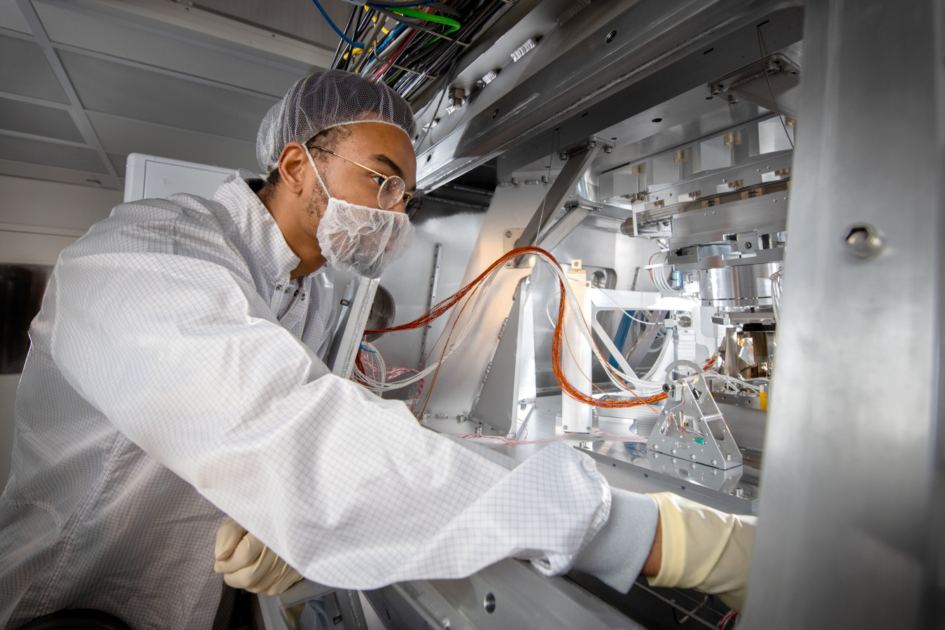 A man in a clean room suit is looking at a complex piece of machinery within a metal cabinet.