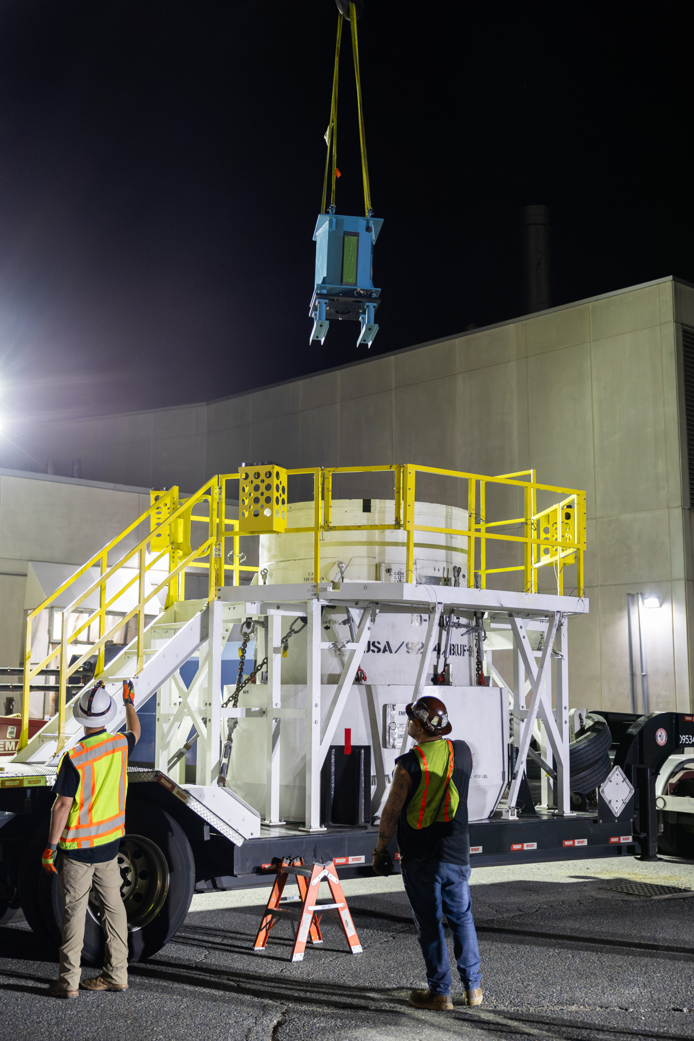 A blue, dishwasher-sized metal container is lowered into a large white, metal cask by crane. There is a yellow staircase surrounding the cask, which is on a semi trailer. In the foreground, men in high-visibility clothing direct the crane operator.
