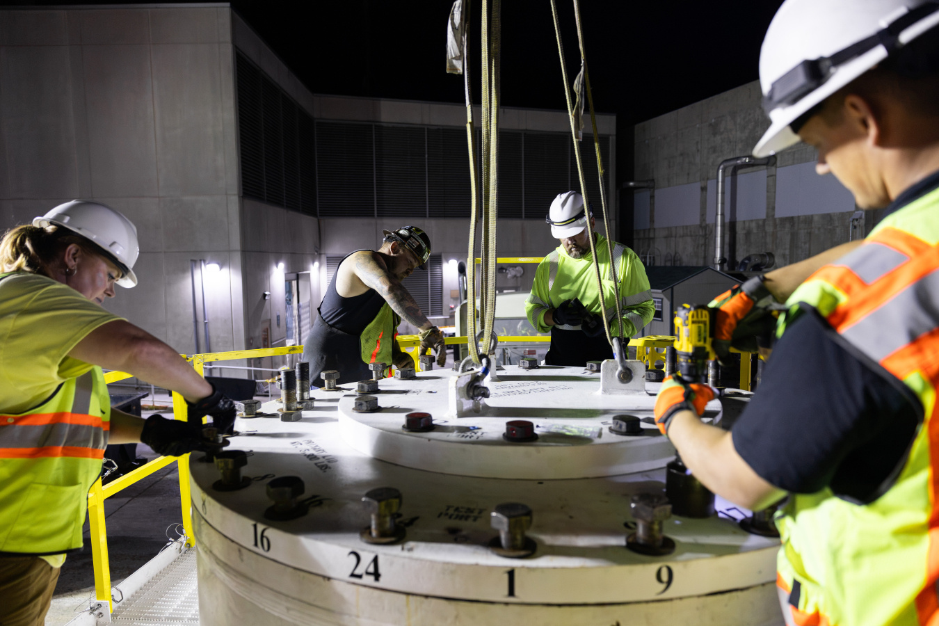 Four men bolt on the top of the metal cask containing the irradiator. They are weaing high-visibility vests and hard hats. The scene is at night and it is eerily lit.