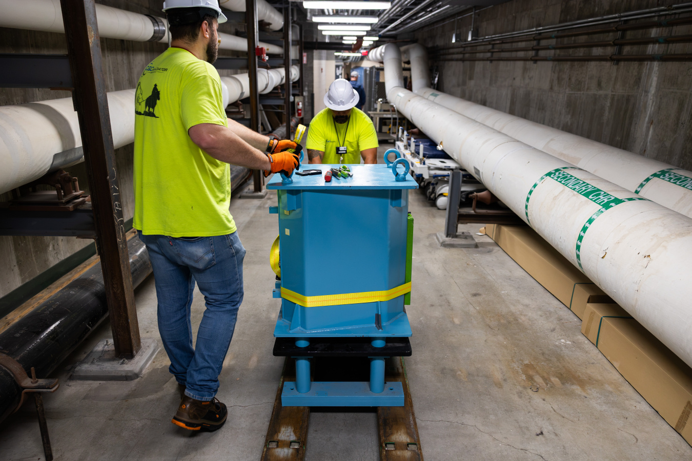 Two men wearing high-visibility vests and hard hats wheel a blue, metal case about the size of a dishwasher down what appears to be a basement corridor of a building. Large pipes are on their left and right.