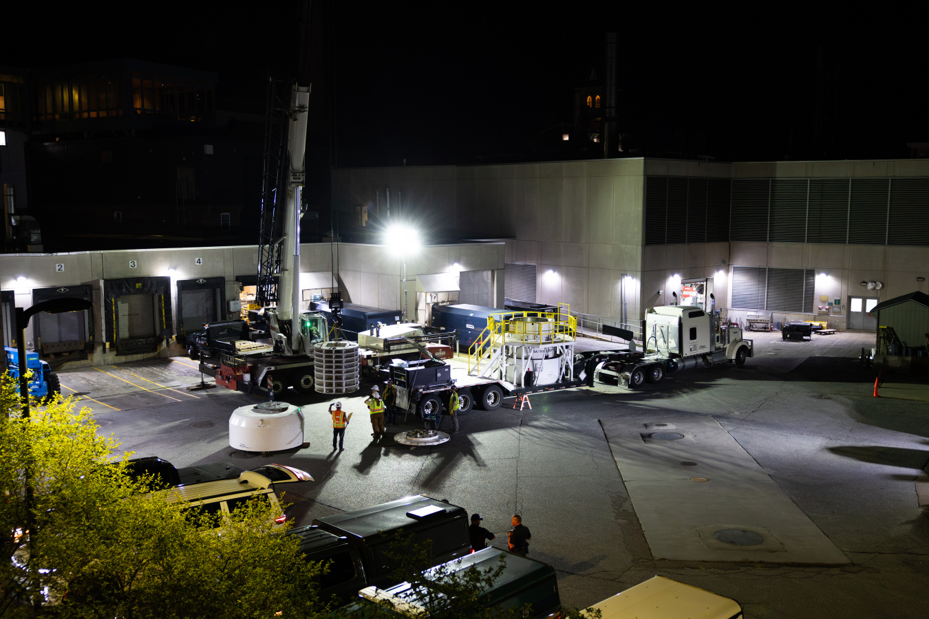 An overall look at the scene of the removal: A parking lot is ablaze with lights as a lone worker in high-visibility clothing and a hard hat directs a crane operator where to put the giant cask lid. 