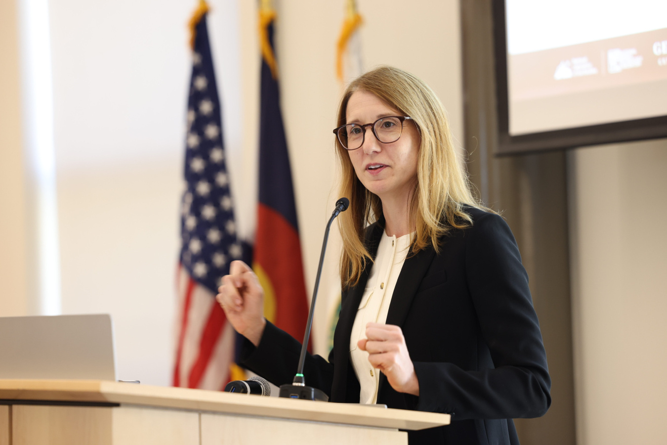 Photo of a woman in a suit and glasses presenting at a lectern with a microphone, with several flags and the corner of a presentation screen in the background.