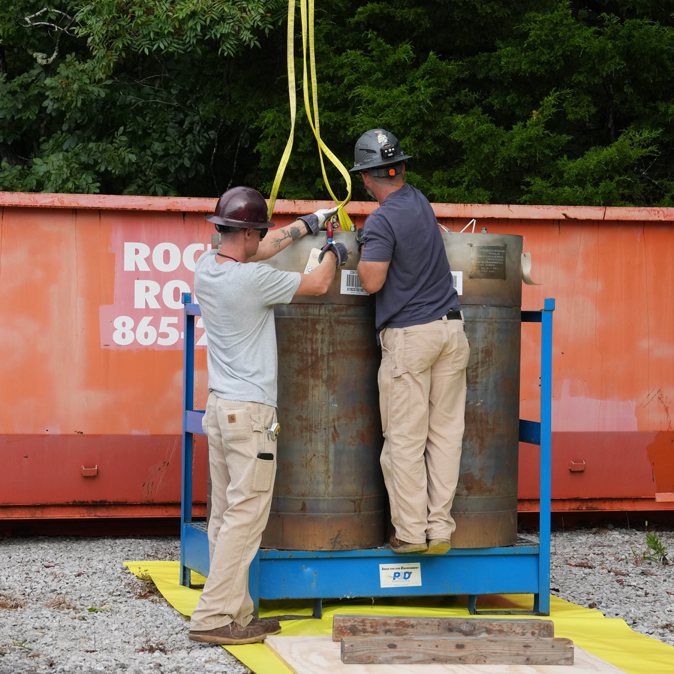 Two workers wearing hard hats attach yellow straps to a pair of large metal cylinders