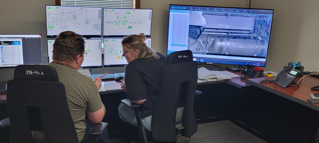Two employees sitting a desk with multiple monitors in front of them, speaking with each other