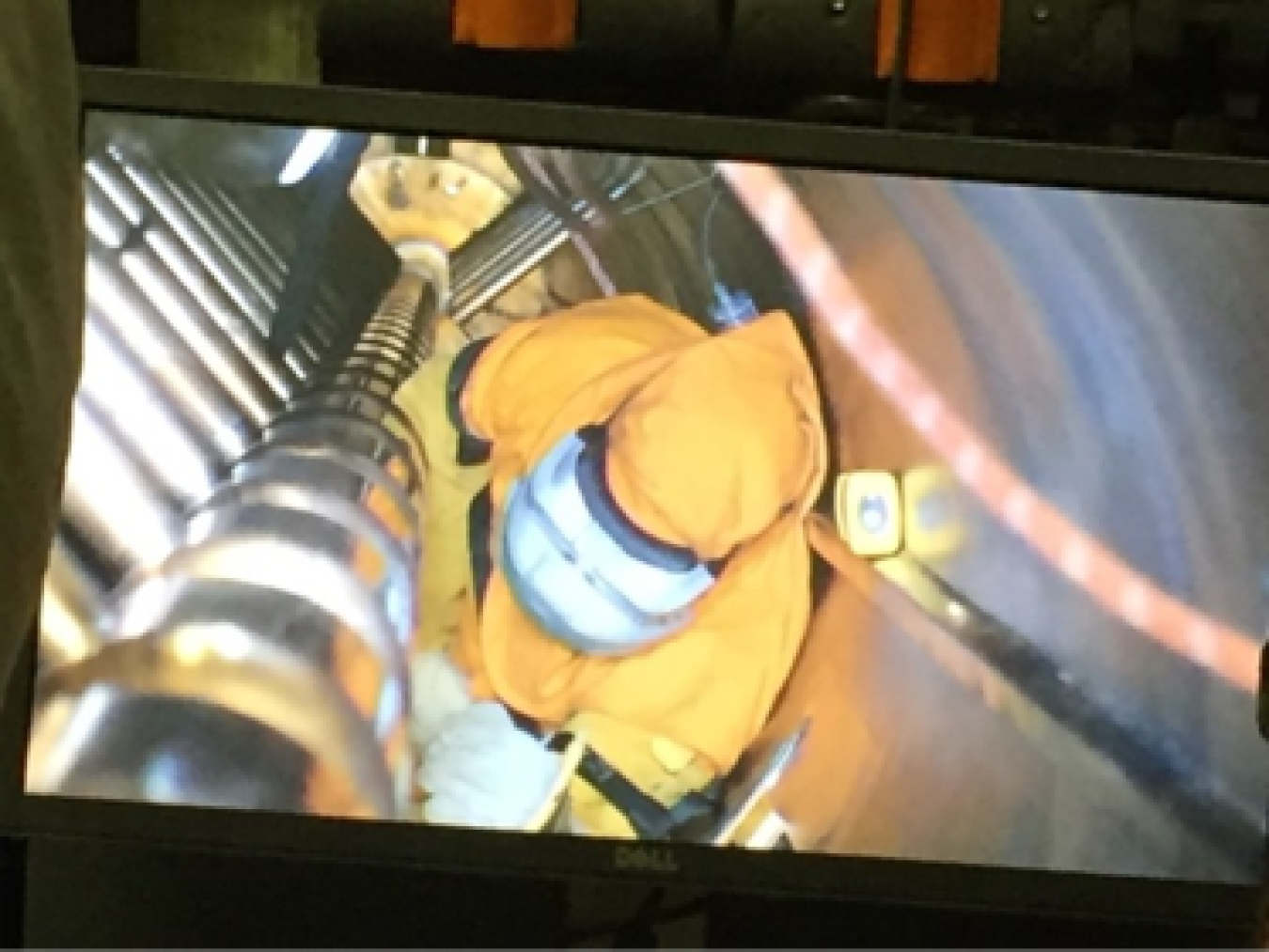 A video image of a man in a protective yellow suit wearing a welder's mask working in a tightly confined, long space.