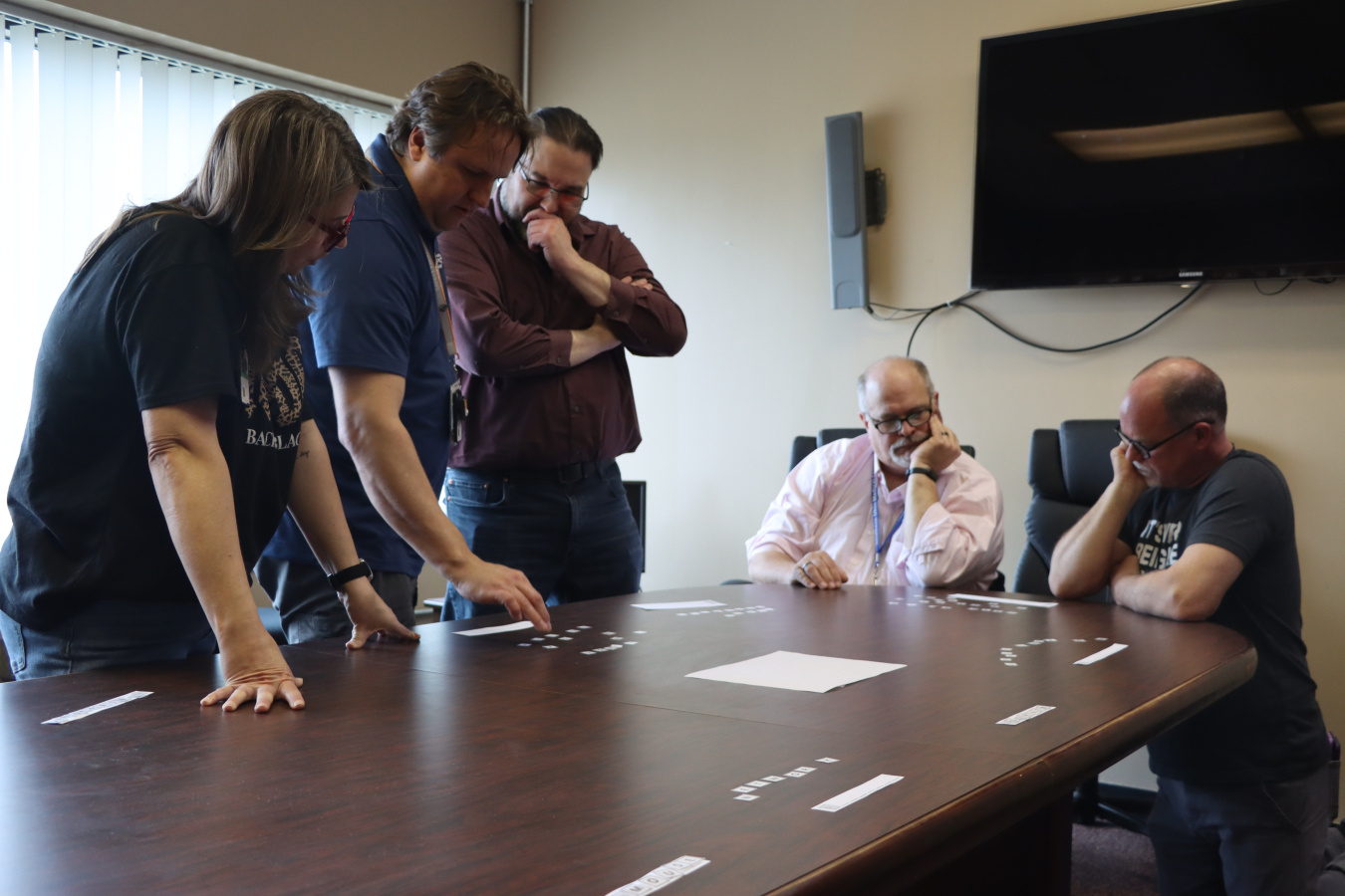 Three employees standing and two sitting looking at a piece of paper on a table