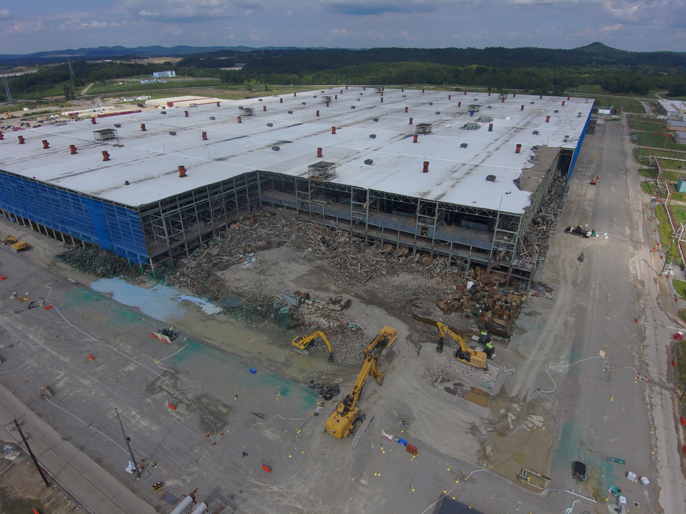 Aerial view of demolition taking place on the corner of a large building with a white roof and blue walls