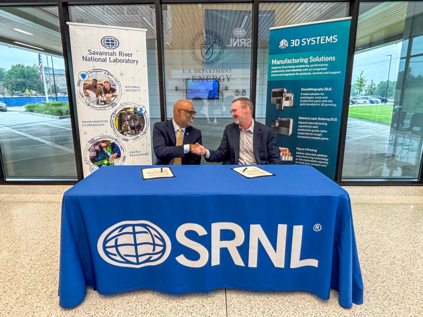 A pair of professionals sitting at a table with a blue tablecloth, they face each other and shake hands