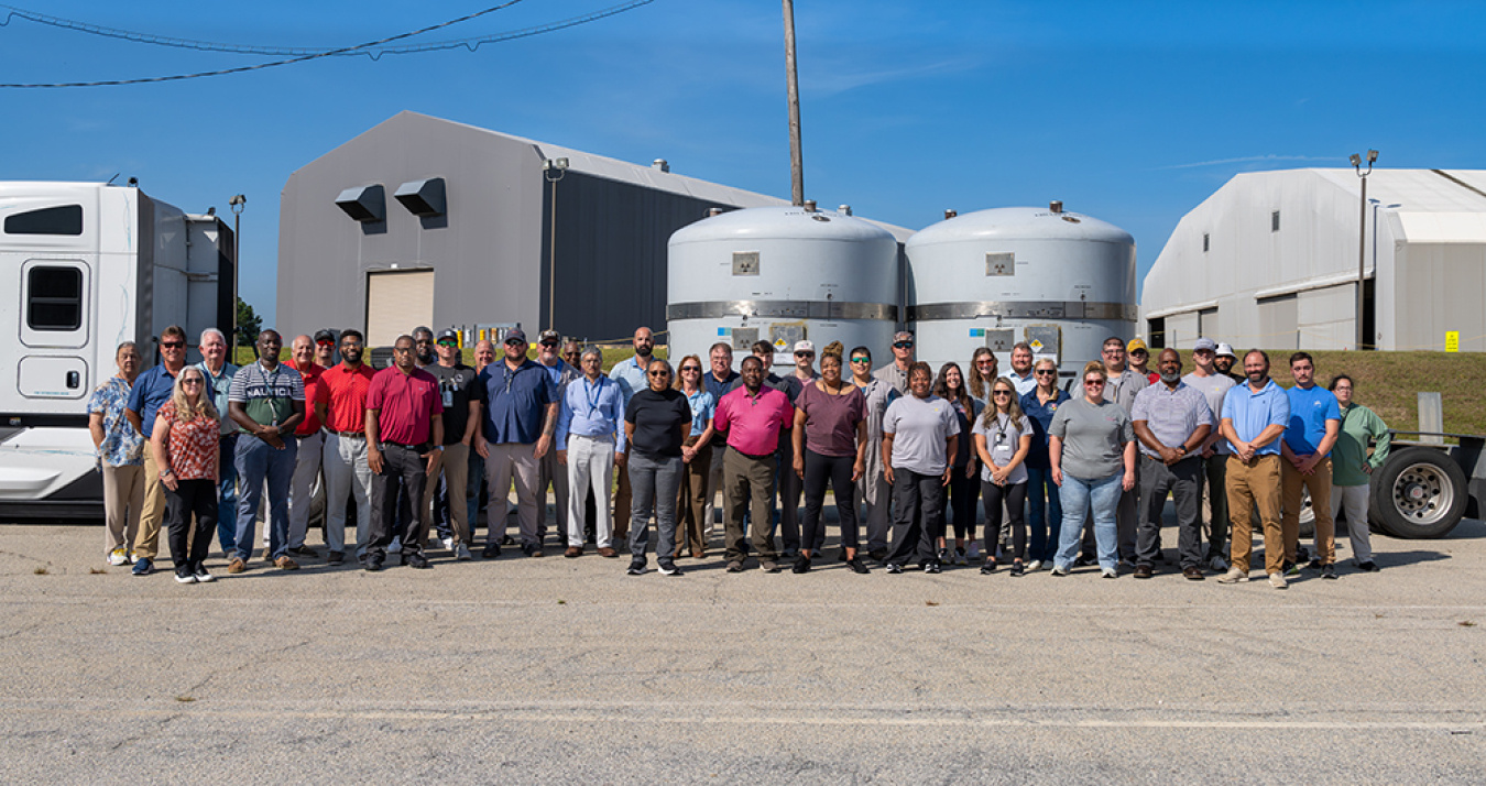 A large group of people posing for a photo outside of a tractor trailer truck with 2 large tanks on the back