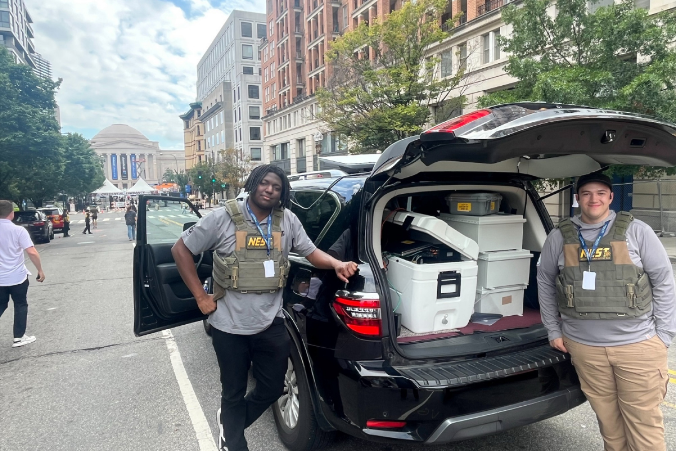 Two people wearing NEST vests stand in front of a packed SUV in an urban environment. The National Mall is behind them.