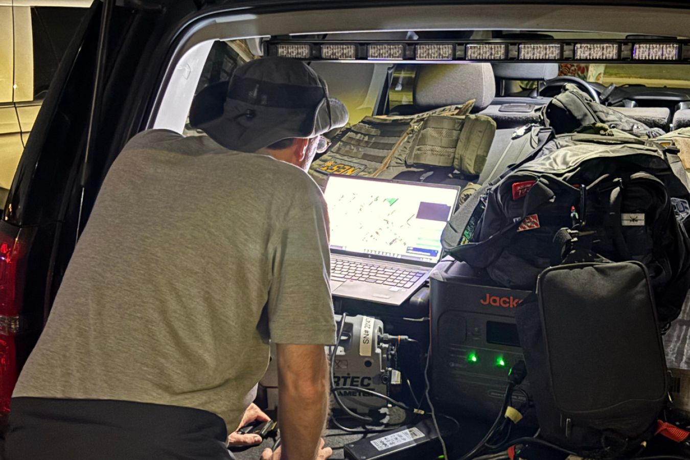 A man monitors a computer screen with a map on it. It's in the back of a well-lit SUV along with a lot of equipment.
