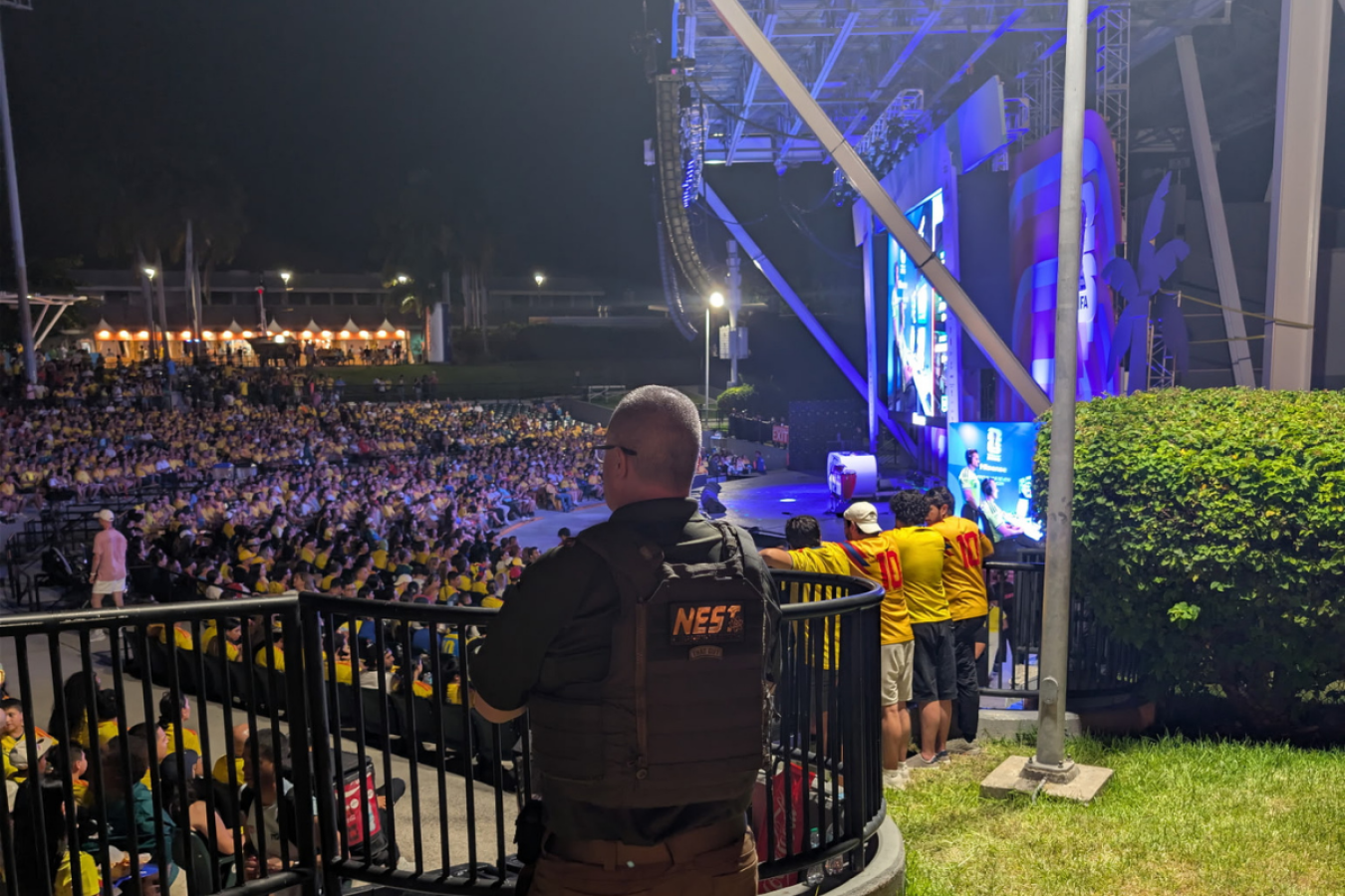 A man wearing a NEST vest watches a raucous crowd of people in front of a large, outdoor TV screen