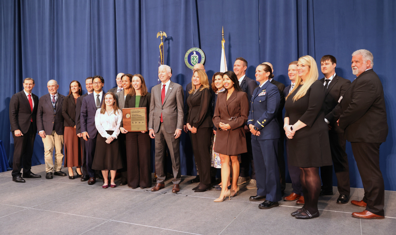 A large group of professionals pose for a photo with the Secretary of Energy, one person holds and award