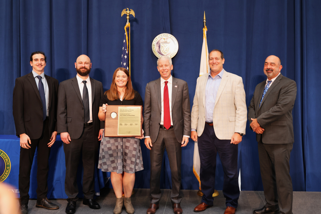 Six professionals pose for a photo with the Secretary of Energy, one person holds an award