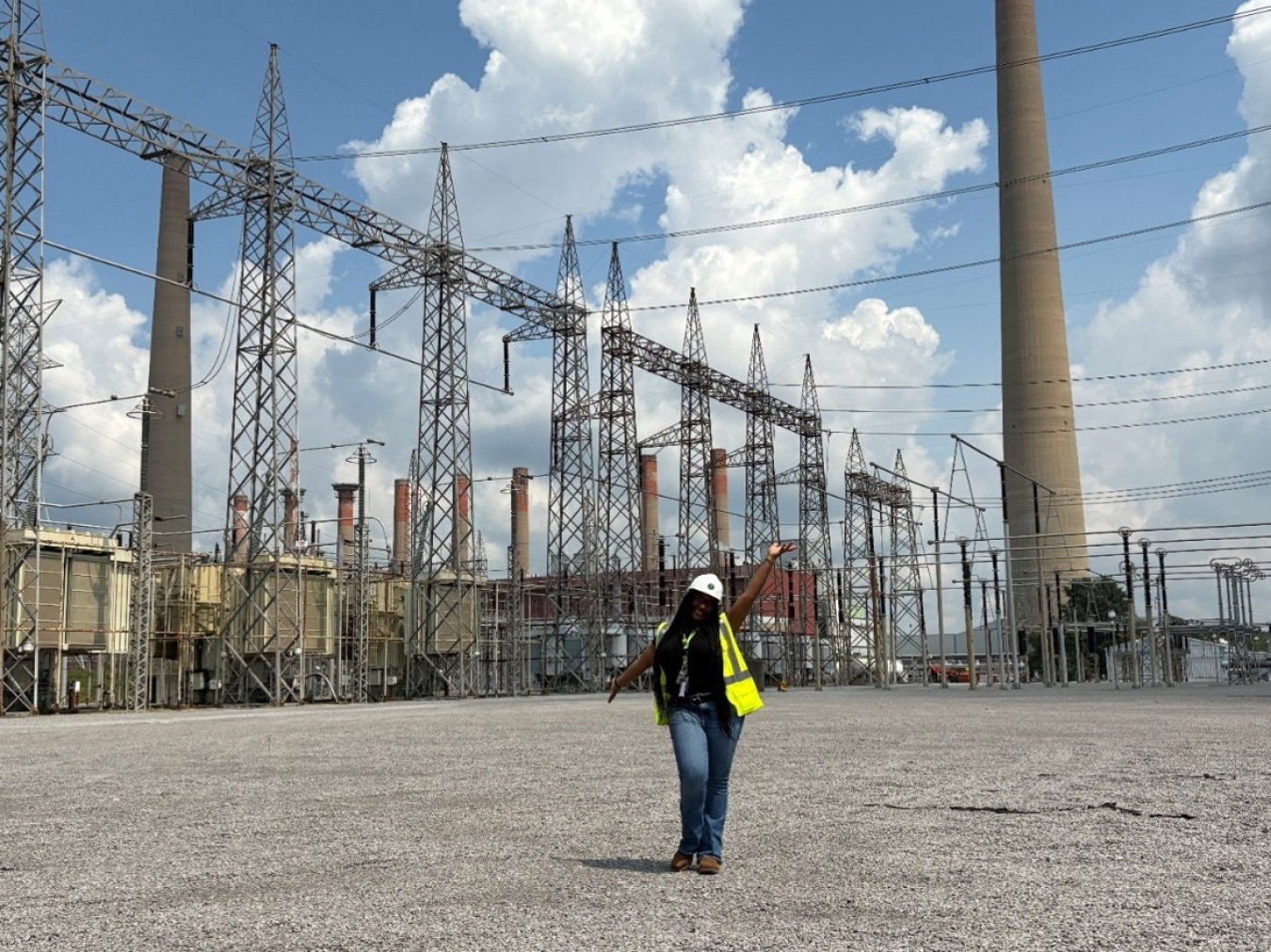 A woman in a white safety hat and yellow safety vest poses for a photo in front of tall electrical towers
