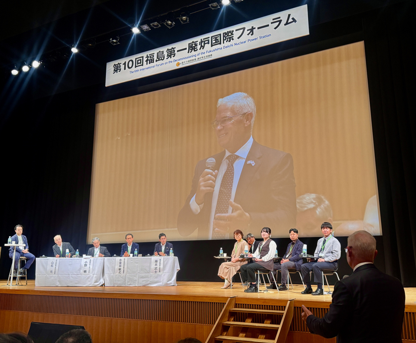 A man on a large screen on a stage speaking to the audience and people sitting in chairs holding microphones