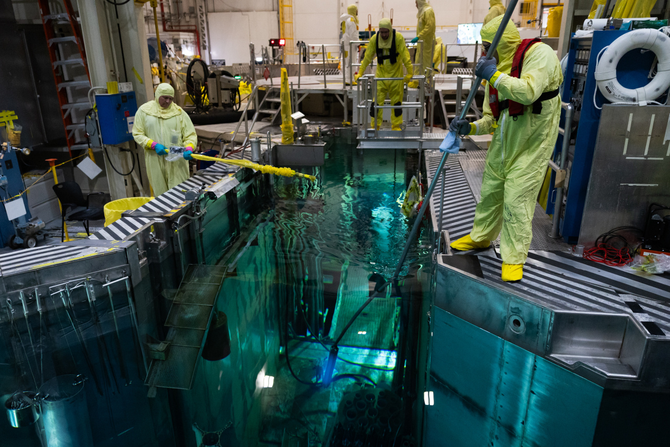 Workers in hazmat suits stand next to a water-filled channel containing nuclear fuel rods.