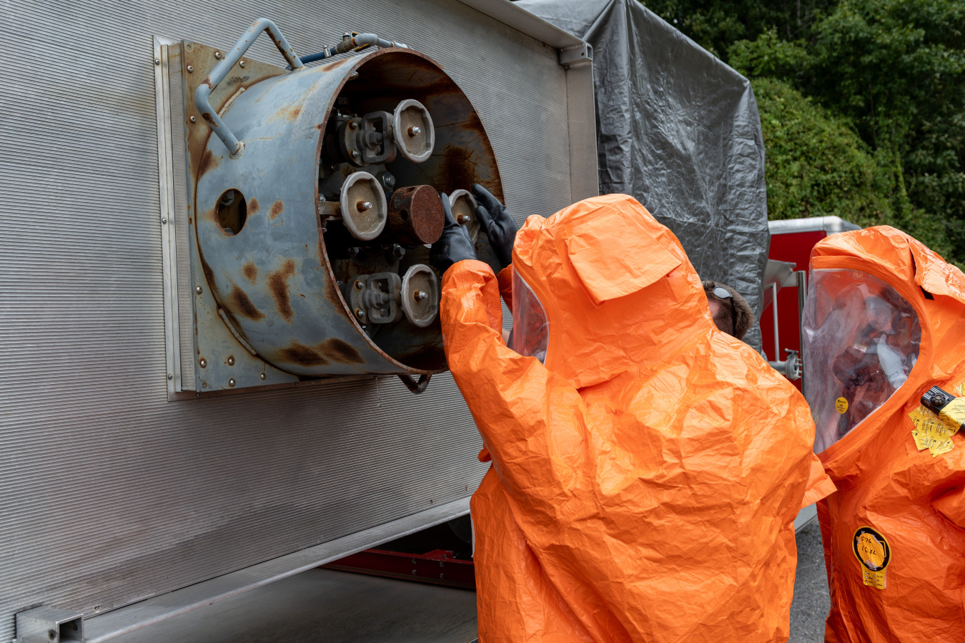Employees in orange hazmat suits secure a leak from a large valve on the side of a wall
