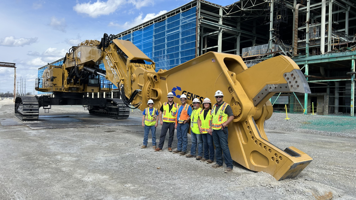 A group of people in safety gear standing beside a large yellow set of excavator shears