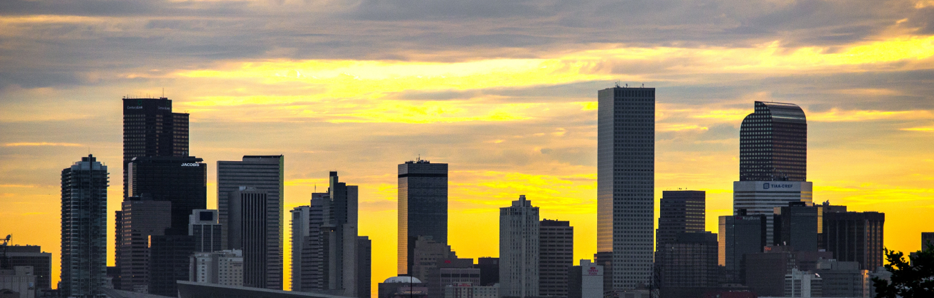 Photo of a city skyline with tall office buildings and a multicolored sky beyond.