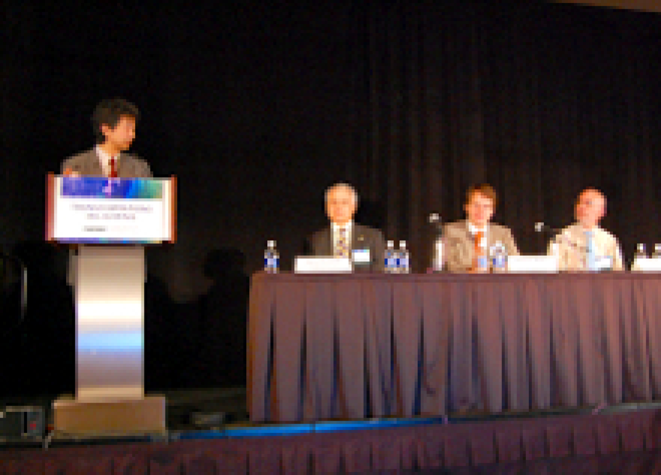 Photo of a man standing at a podium, with three people seated at a table to his left.