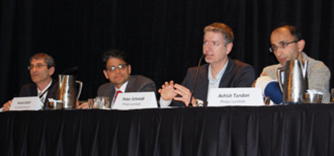 Four men sitting at a long table as panelists, addressing the audience via microphones.