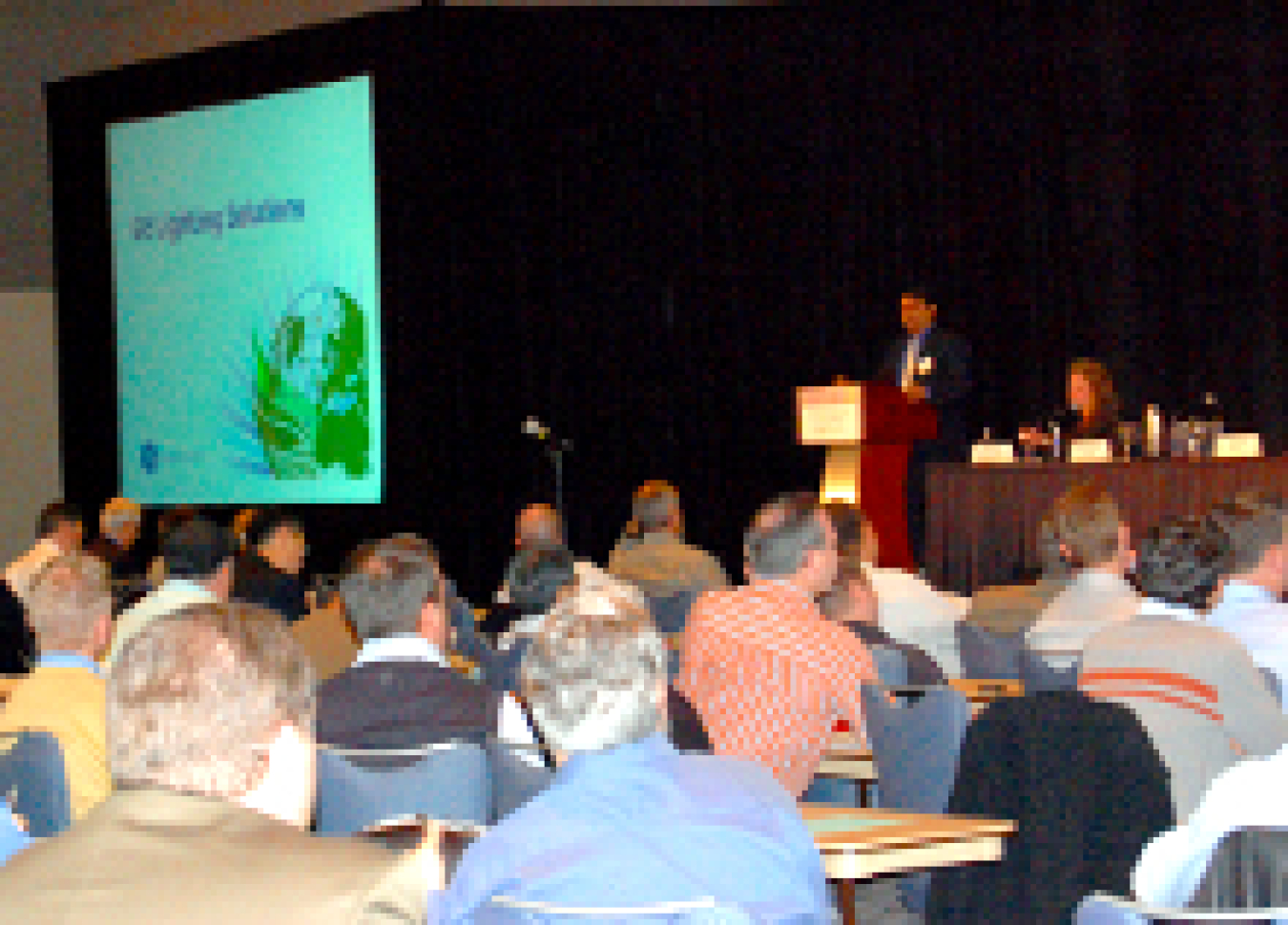 Photo of a group of people assembled in a room, with their backs to the camera, seated and listening to a speaker.