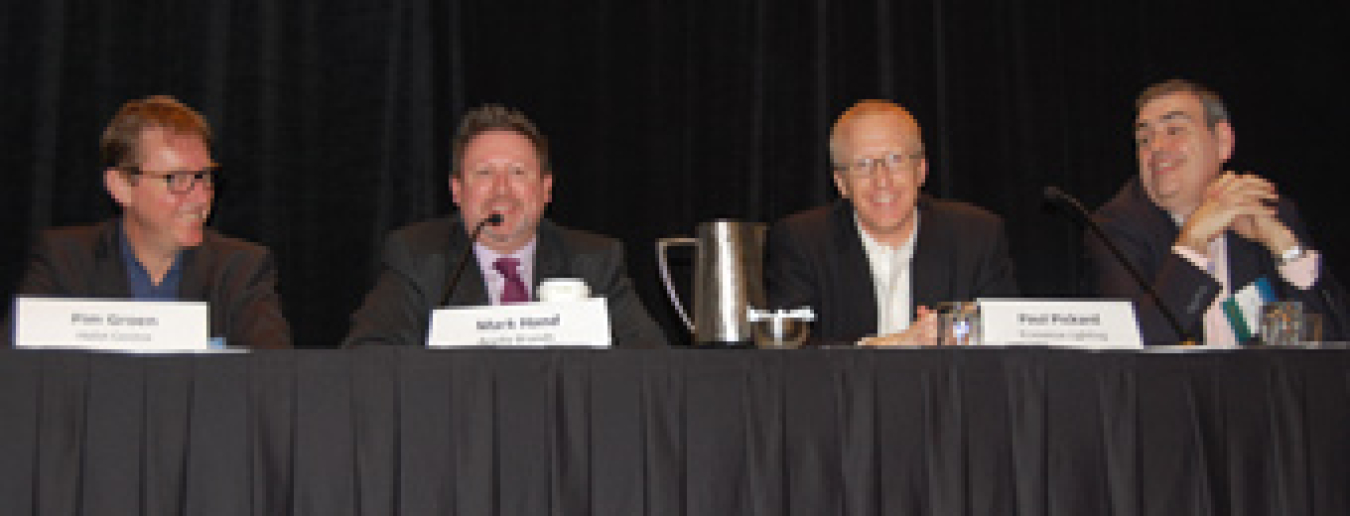 Four men sitting at a long table as panelists, addressing the audience via microphones.