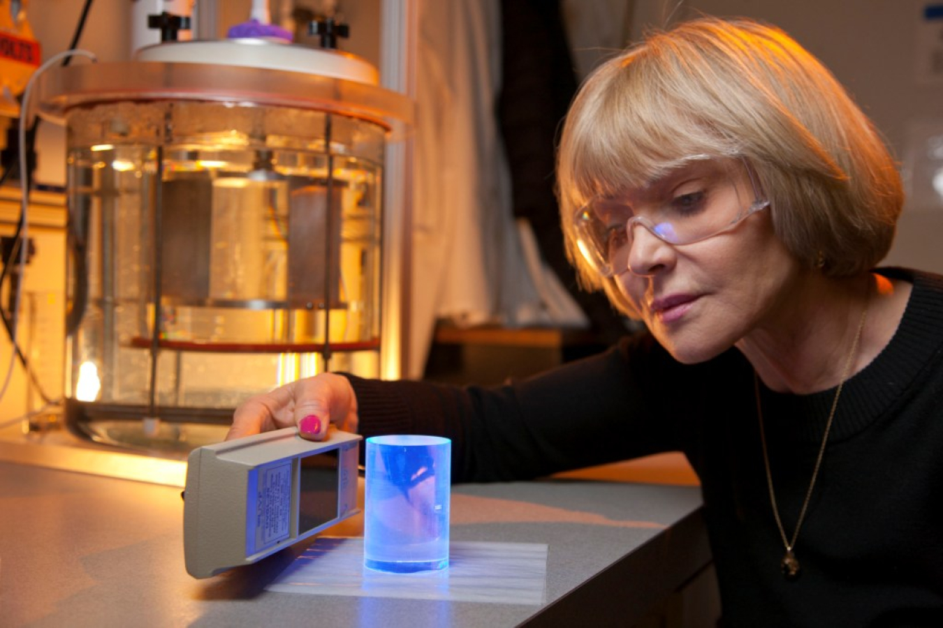 Woman wearing safety glasses, with a handheld instrument, examining a small container in a laboratory.