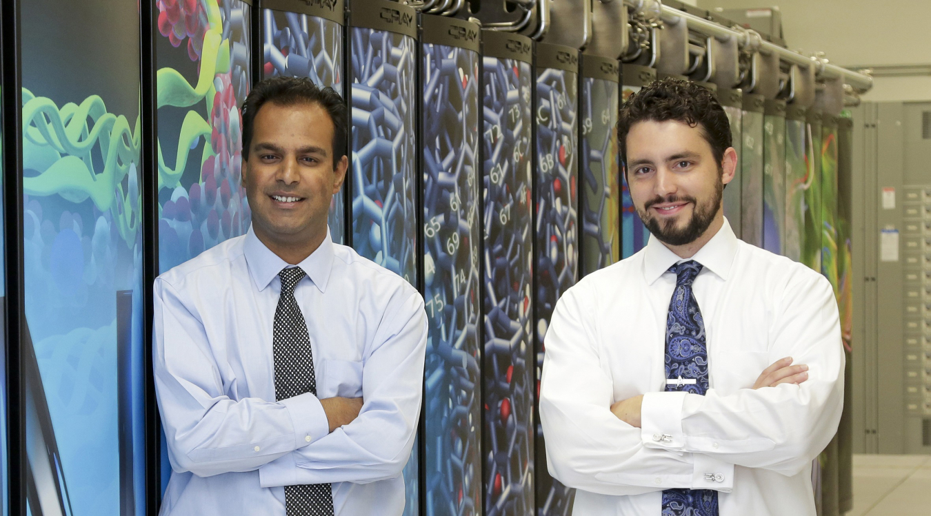 Photo of two men standing, facing the camera, in front of a decorative wall.