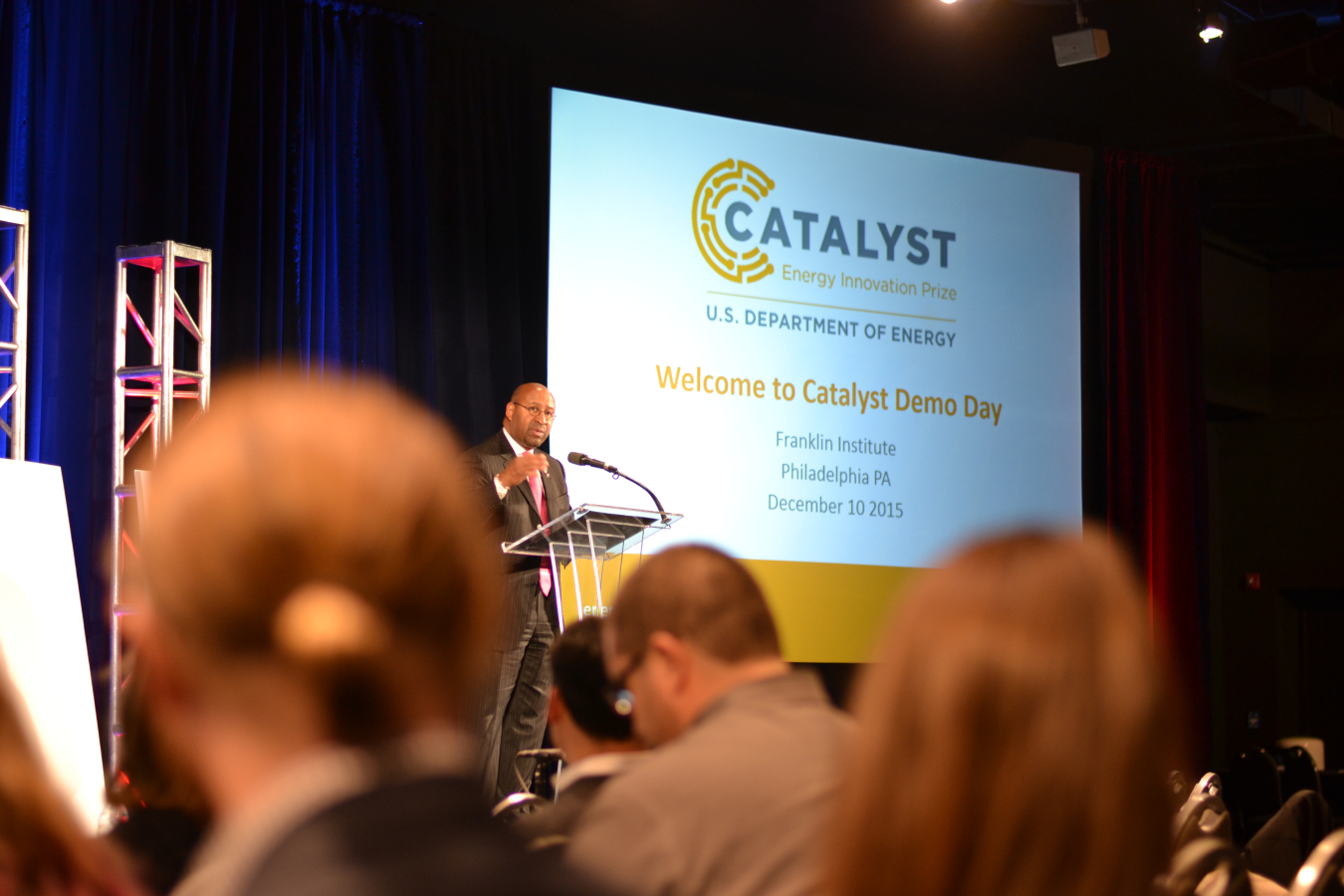 Audience perspective / view of a speaker at a lectern, with a projection screen to the right, addressing the group.