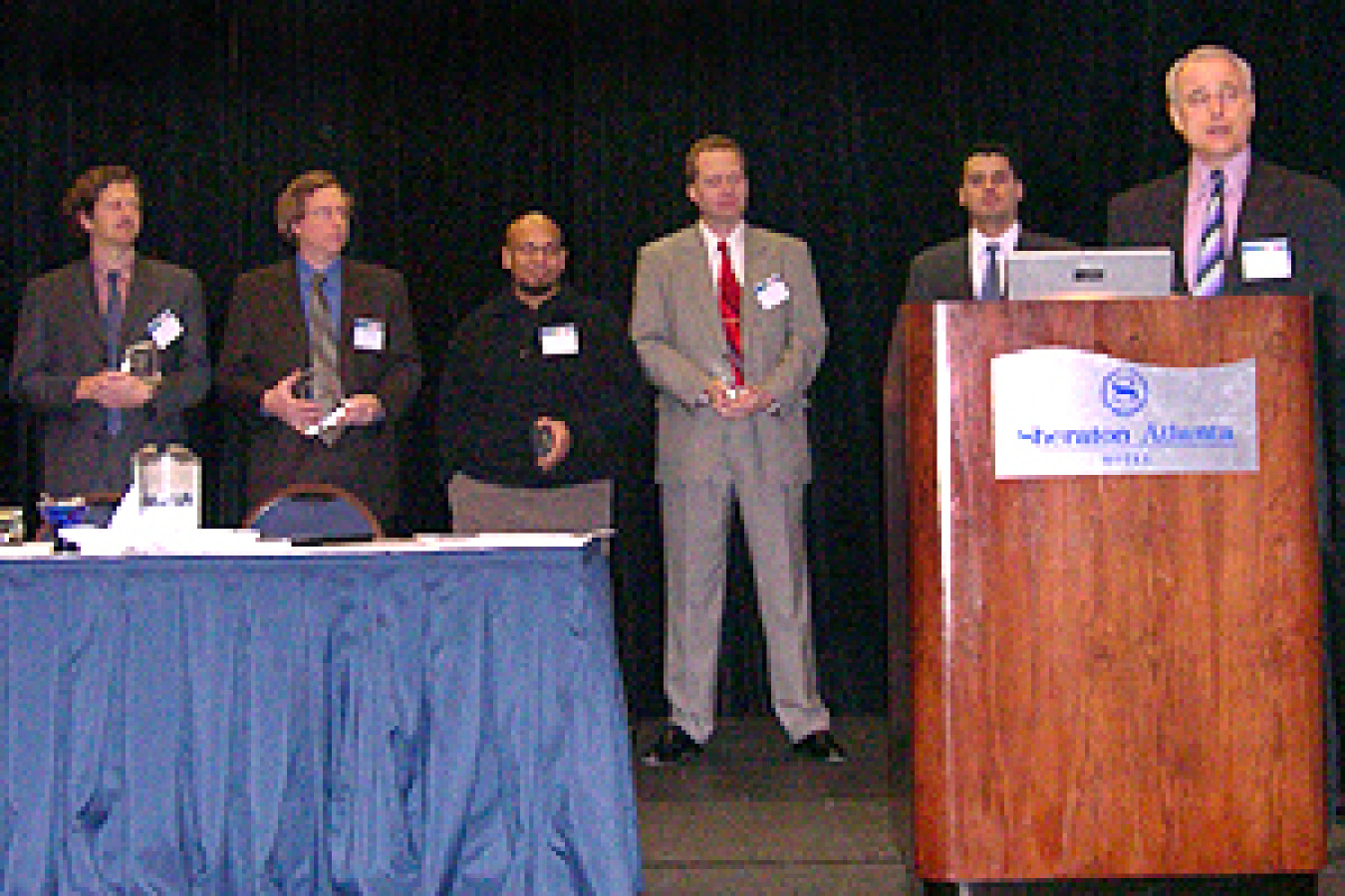 Photo of 5 men standing behind a long table, with a sixth man standing behind a podium, which is on the right.