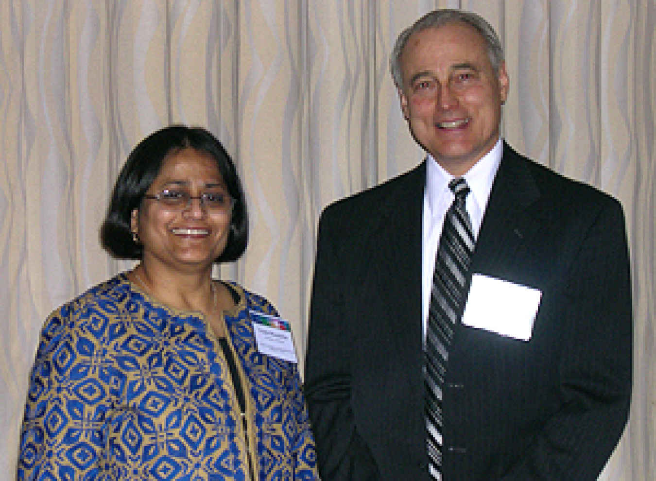 Photo of a man and a woman, standing, smiling at the camera. The woman, on the left, is wearing a colorful blue and gray outfit. The man, on the right, is wearing a suit.