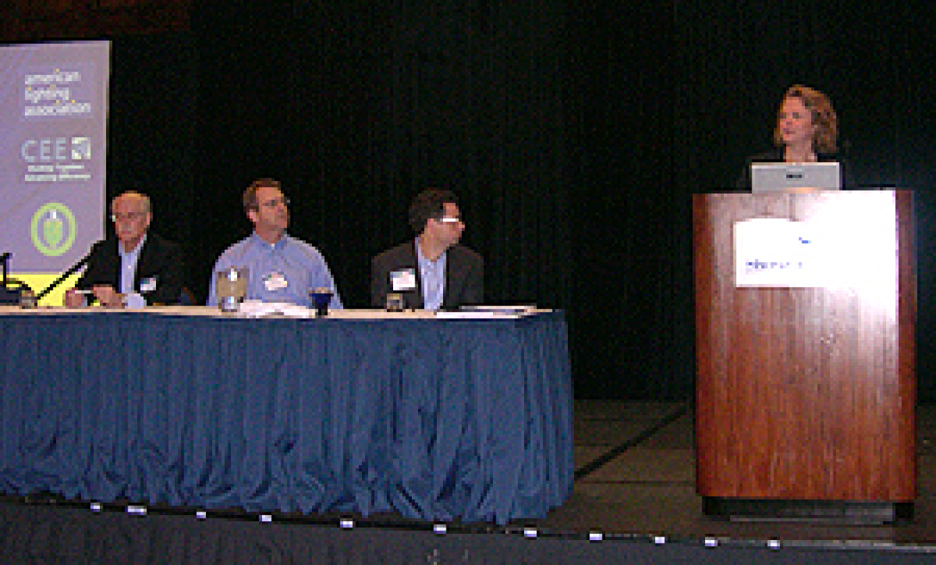 Photo of three men seated behind a long table. Two of the men are looking to their left at a woman standing behind a podium.